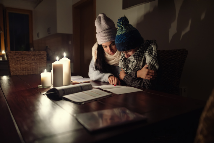 Shown is a colour photograph of people in a dark room wearing winter clothes inside and reading books by candlelight.